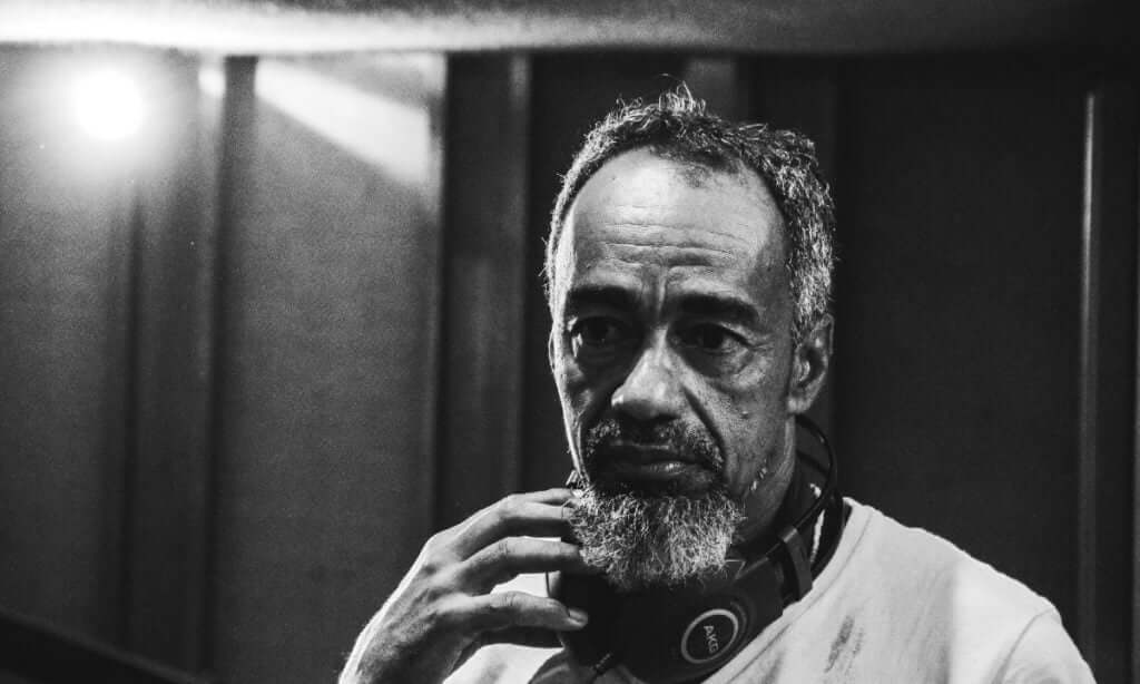 Foto de Ubiratan Marques, maestro da Orquestra Afrosinfônica da Bahia. Homem negro, 50+, com barbicha e cabelo grisalho, usando um fone de ouvido no pescoço