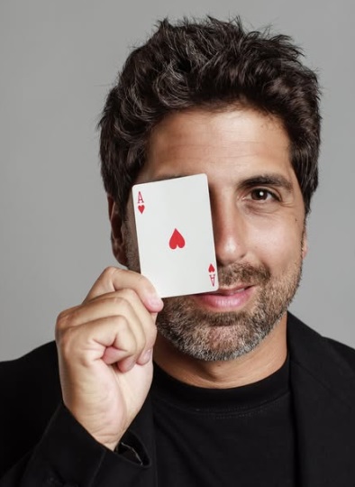 O carioca Gabriel Louchard está segurando uma carta de baralho a frente do rosto. Ele é ator, comediante, apresentador e ilusionista.